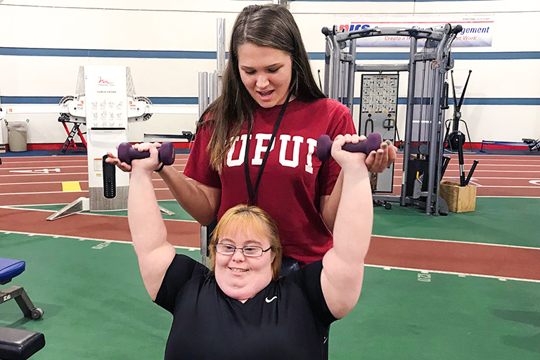 A female AMP instructor in an IUPUI sweatshirt spots a female AMP client lifting dumbbells.