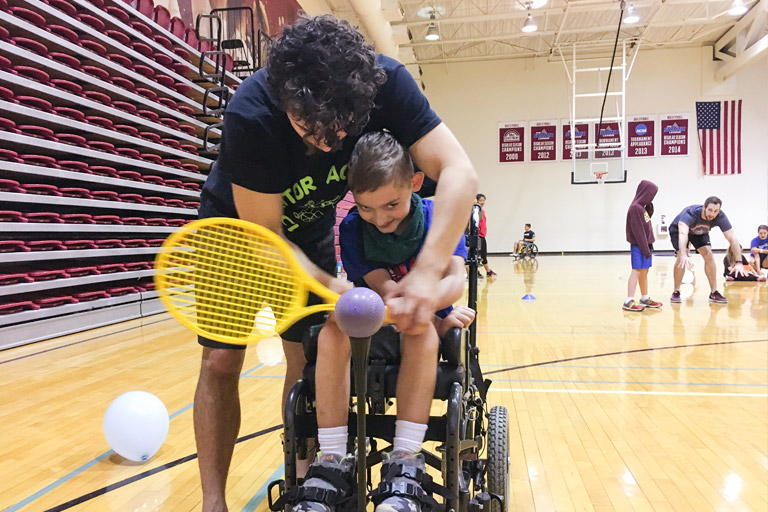 An AMP instructor helps a little boy in a wheelchair hit a tennis ball with a racquet.